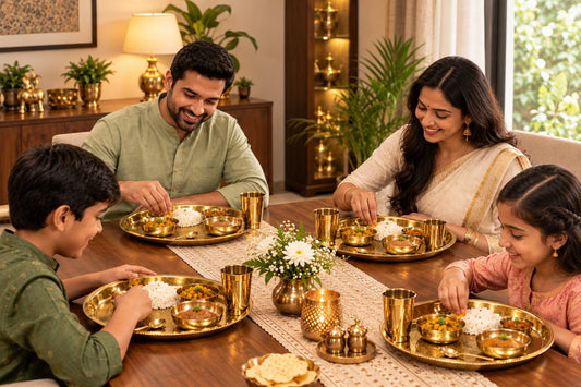 A family of four sharing a meal together around a wooden dining table, using traditional brass Indian thali plates with rice, samosas, and small brass cups in a warmly lit home setting.