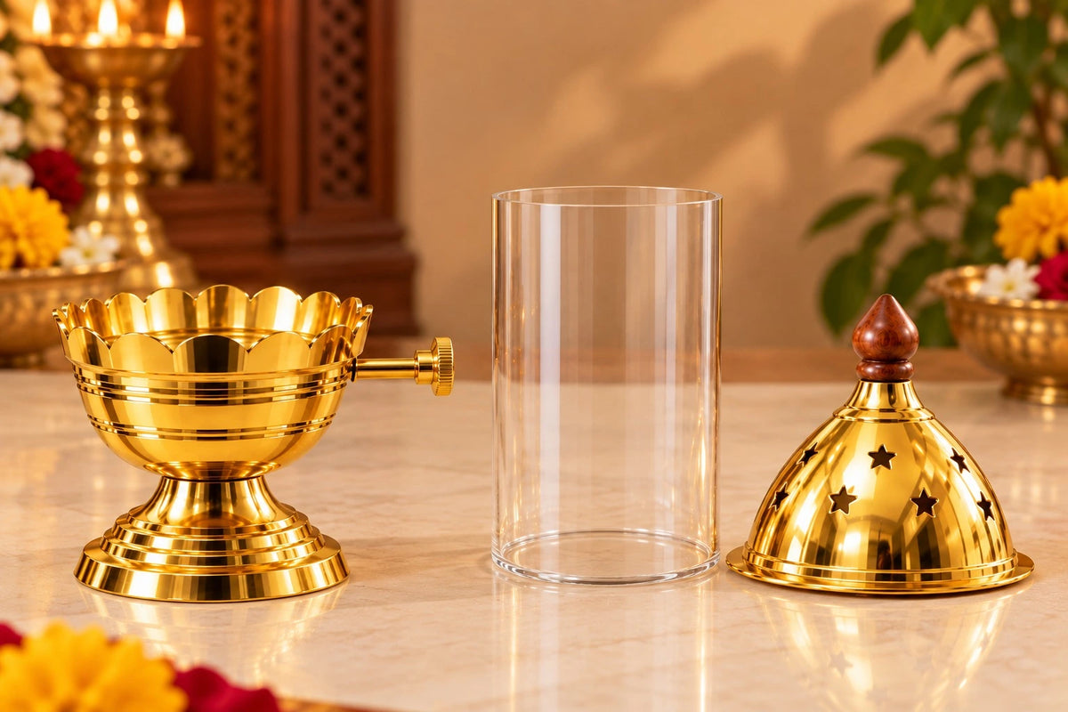 A brass oil lamp set with a gold-toned lamp base, a clear cylindrical glass jar, and a matching gold-toned lamp cover with star cutouts, displayed on a marble tabletop with warm indoor background lighting. Akand Jyot diya