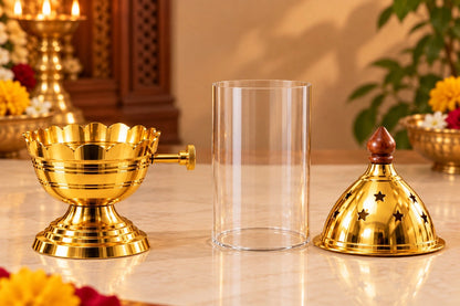A brass oil lamp set with a gold-toned lamp base, a clear cylindrical glass jar, and a matching gold-toned lamp cover with star cutouts, displayed on a marble tabletop with warm indoor background lighting. Akand Jyot diya
