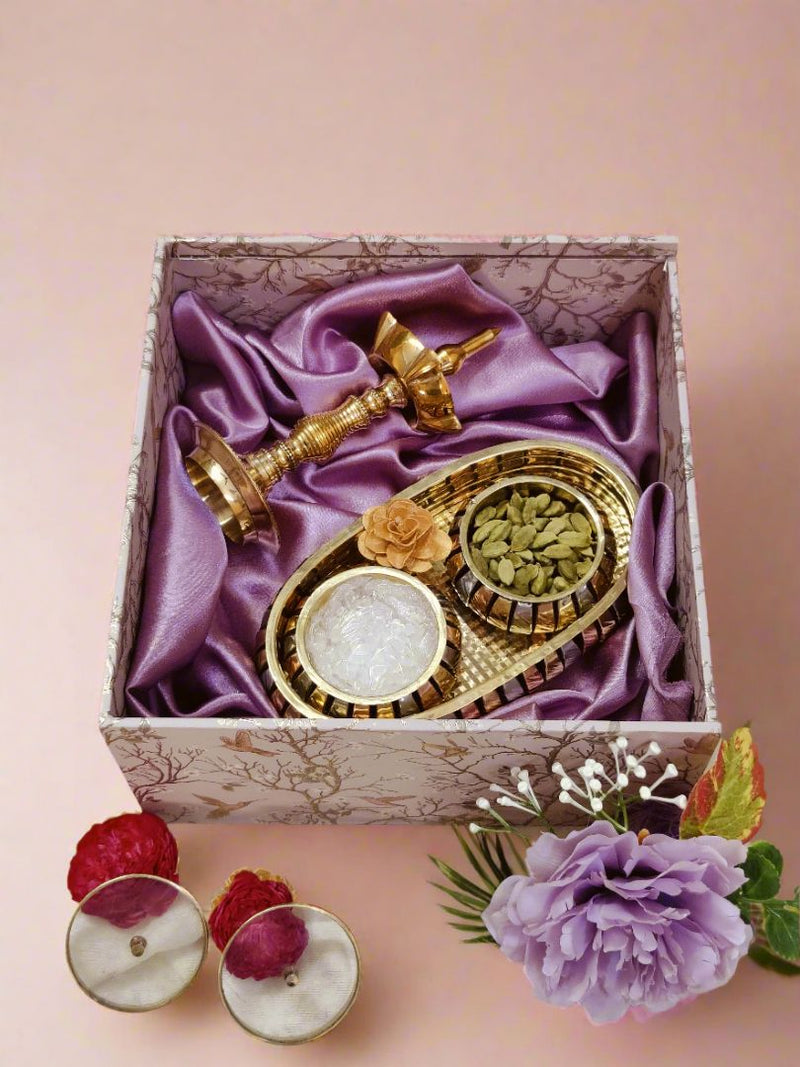 Aroma & aura gift box featuring a brass tray with cardamom and sugar crystals 
beside a brass diya, arranged in a purple silk-lined premium brass gift box.​