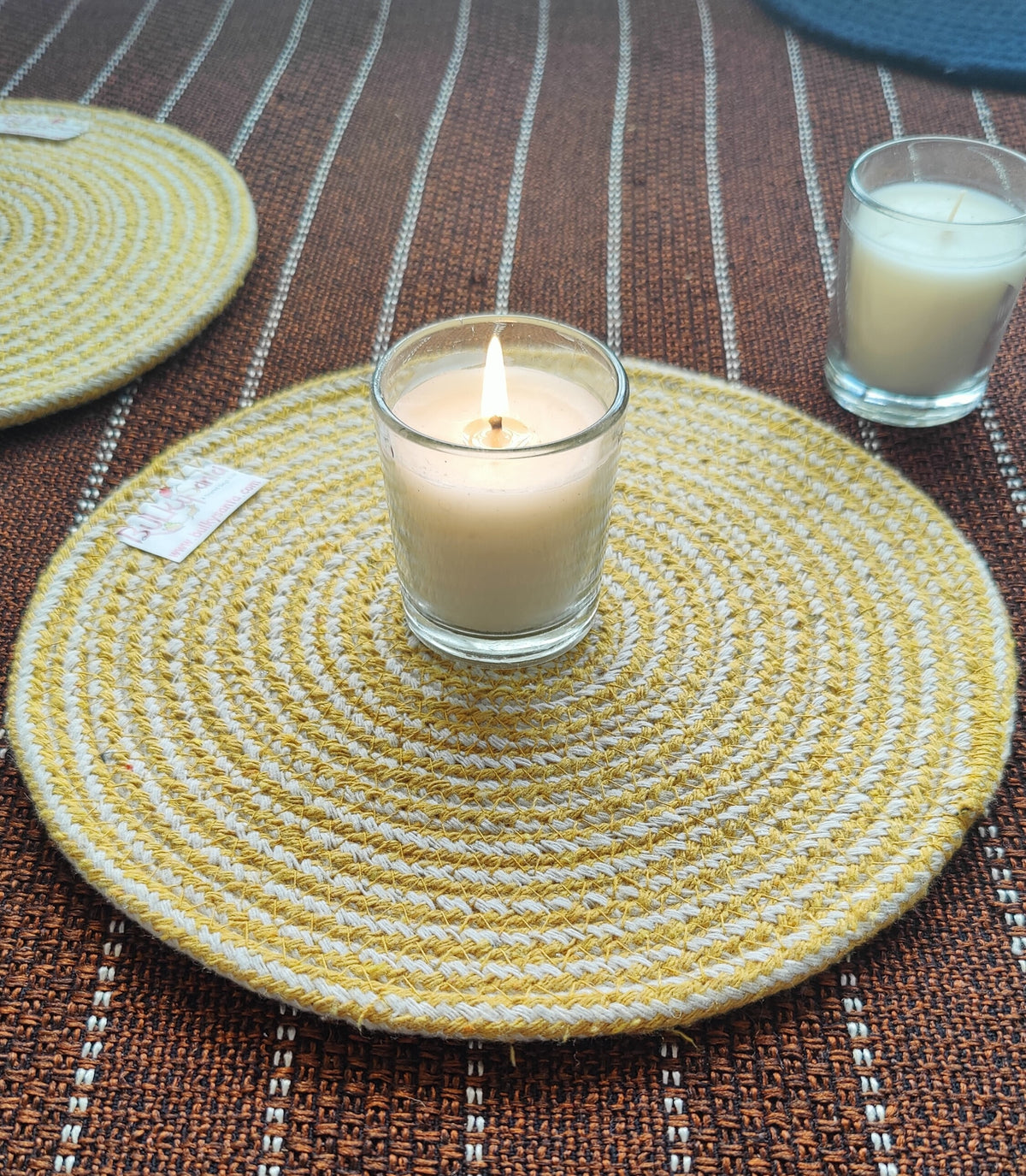 Candle in a glass holder on a woven yellow coaster with a striped brown tablecloth.