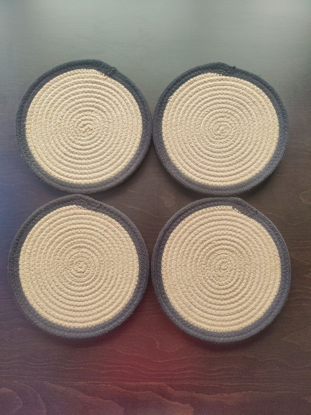 Four round woven coasters with beige center and dark gray border on a wooden surface.