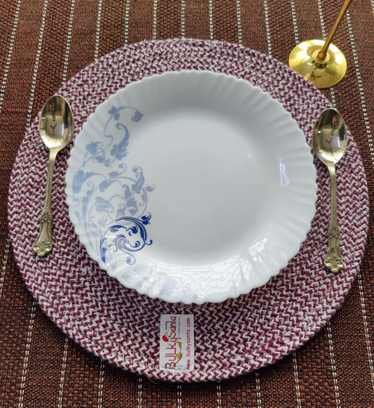 Close-up of a maroon woven round placemat with a floral white plate and spoon arranged for dining.