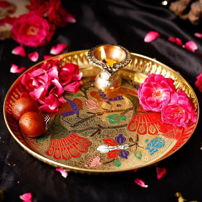 Decorative plate with floral design, lit candle, and pink flowers on a dark background peacock design thali brass